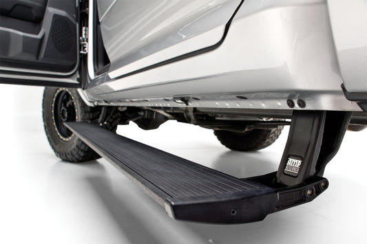 Close-up of an AMP Research 22-23 Chevy/GMC Silverado/Sierra 1500 & 2024 2500/3500HD PowerStep Plug N Play running board, extended from a silver vehicle. The board showcases a textured black surface and is firmly secured with a robust bracket, accentuating details of the vehicle's undercarriage.
