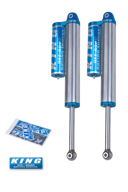 The image features a pair of King Shocks, specifically the 2011+ Chevrolet Silverado 2500/3500 Rear 2.5 Dia Piggyback Reservoir Shocks, in silver and blue. These shocks, recognized for exceptional ride quality with their piggyback reservoirs, come with a set of King decals and one large blue sticker, providing enhanced shock absorption for challenging terrains.