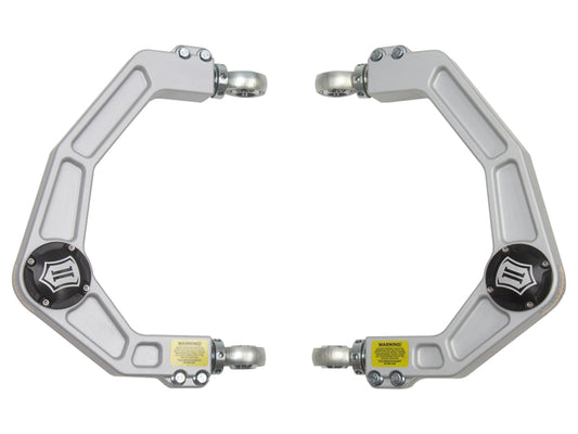 The ICON 2021+ Ford F-150 Billet Upper Control Arm Delta Joint Kit from ICON showcases a Delta Joint design for improved flexibility, a silver-gray finish, and black circular center components. Each metal arm is equipped with mounting brackets at both ends that have yellow warning labels, ensuring they are resistant to corrosion and symmetrically oriented.