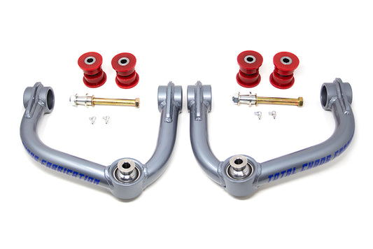 The TOTAL CHAOS 21-23 Ford F-150 Upper Control Arms, featuring gray suspension arms with red bushings and bolts, are arranged symmetrically on a white background. The upper control arms, marked with blue text, are crafted to enhance performance in aftermarket lift kits.