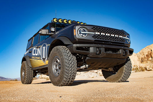 A rugged, black off-road vehicle with "Bronco" on the grille and "ICON" on the side is parked on sandy terrain. Equipped with ICON's 21-23 Ford Bronco 2.5in VS RR Coilover Kit - Front, it features large tires, raised suspension for enhanced performance, and extra roof lights.