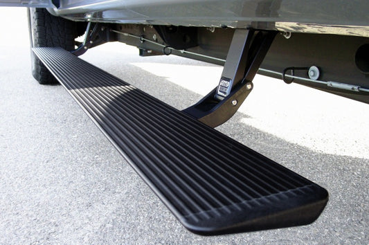 The AMP Research 1999-2006 Chevy/GMC Silverado/Sierra Extended/Crew PowerStep in black is depicted in a close-up shot, with its ribbed, electric-powered running board extended and installed beneath the vehicle. The automatic running board is displayed in profile against a grey pavement background.