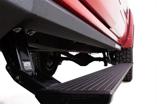 Close-up view of an AMP Research 2014-2017 Chevrolet Silverado 1500 Crew Cab PowerStep XL in black, mounted beneath a red lifted truck. The retractable step is extended, highlighting its textured surface and robust construction, with the vehicle's tire and suspension visible in the background.
