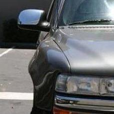Close-up of the front right corner of a gray vehicle, showing part of the hood, front grille, headlight, and side mirror. The car is parked in a parking lot and is equipped with ADV Fiberglass 1999-2002 Chevrolet Silverado fenders. The background includes pavement and indistinct objects.