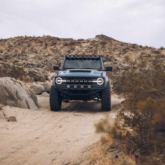 A rugged Ford Bronco with off-road modifications, including large tires, an ADV Fiberglass 2021-2023 Ford Bronco Ram Air Hood, and a roof rack, is parked on a sandy trail surrounded by rocky desert terrain under an overcast sky. Shrubs and boulders dot the arid landscape.