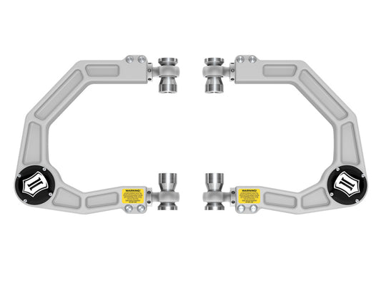 These ICON 2021+ Ford Bronco Billet Upper Control Arm Delta Joint Kit brackets are essential suspension components designed for off-road performance. Crafted from silver metal and featuring symmetrical designs with geometric patterns and visible bolts, each bracket is marked by a black circular label showcasing a white emblem, along with yellow warning labels. The mirrored Delta Joint PRO brackets are positioned horizontally facing each other.