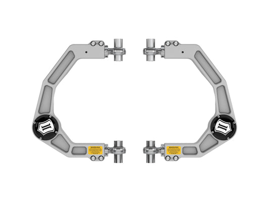A pair of silver, geometric mechanical clamps are symmetrically arranged on a white background. These clamps, inspired by the design of the ICON DJ Pro Kit from the ICON 21-23 Ford F150 Billet Front Upper Control Arms, each feature a central black circular component adorned with white logos and small yellow warning labels to enhance off-road performance.