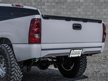 The rear view of a white pickup truck with a lifted suspension and large off-road tires shows impressive ground clearance. The tailgate is closed, and the truck is parked on a gravel surface in front of a brick wall. The license plate reads "MAZZULLA," showcasing the sturdy 1999-2006 GM 1500 REAR PLATE BUMPER crafted by MAZZULLA OFFROAD from CNC cut plate steel for durability.