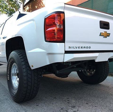 Rear view of a white Chevrolet Silverado pickup truck parked on the street. The Chevrolet logo and the name "Silverado" are visible on the tailgate. The truck is equipped with a 2014-2018 GM 1500 Rear Plate Bumper from MAZZULLA OFFROAD, and it features large tires, impressive ground clearance, and a clean exterior.