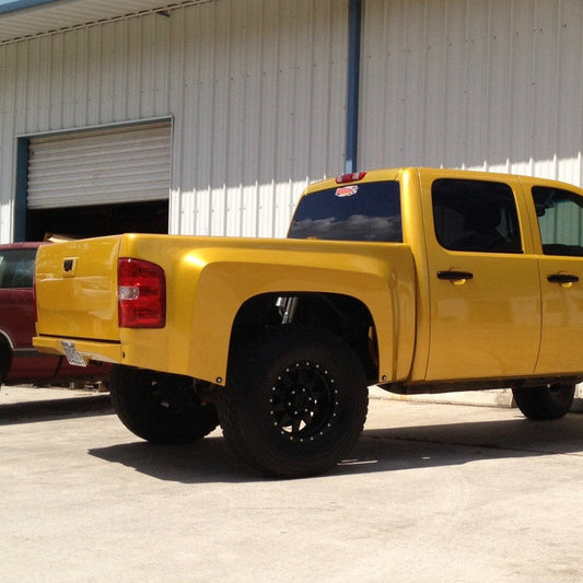 A bright yellow pickup truck with an extended cab is parked beside a red vehicle in front of a large metal building. The truck, enhanced with ADV Fiberglass 2007-2013 Chevrolet Silverado bedsides, boasts large black off-road tires and tinted rear windows. A partially open garage door is visible in the background.