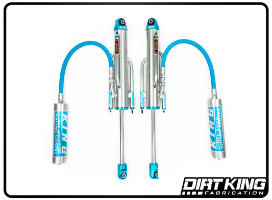 A pair of Dirt King Ford F-150 Long Travel Spec King Triple Bypasses featuring blue hoses. The shocks are sleek and metallic, accented with blue details including the Dirt King logo and text. The DIRT KING SHOCKS logo is displayed at the bottom right of the image. Designed for optimal suspension dampening, the image is framed with a black border.