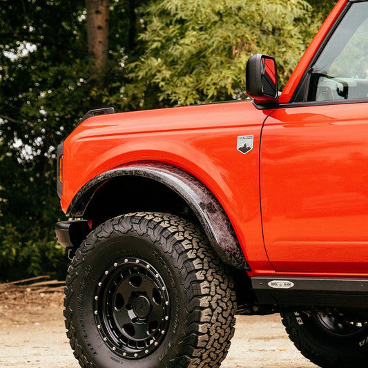 A close-up of the front side of an orange off-road vehicle with large, rugged tires, equipped with ADV Fiberglass's 2021-2023 FORD BRONCO SASQUATCH FLARES. The vehicle features a side mirror with an additional small mirror attached. Trees and greenery are visible in the background.