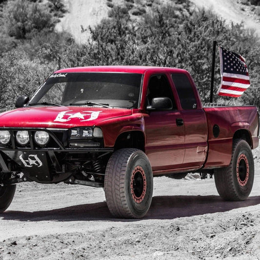 A red off-road truck with large tires and a front grille guard is parked on a dirt road. Equipped with 2000-2006 GMC Sierra Fenders from ADV Fiberglass, it proudly displays an American flag mounted on the back. The background features sparse desert vegetation and rocky terrain.