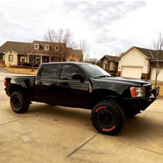 A black 2007-2013 GMC Sierra, equipped with off-road tires and ADV Fiberglass prerunner bedsides, is parked on a residential street. The truck boasts lifted suspension and dark-tinted windows. In the background, there are suburban houses with well-maintained yards and leafless trees, suggesting a cold season.