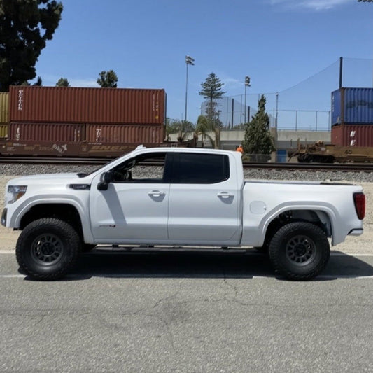 Side view of a white pickup truck with large tires, parked on the side of the road. This 2019-2021 GMC Sierra features sturdy ADV Fiberglass bedsides. Behind the truck, there are several stacked cargo containers and a chain-link fence, with a few trees and light poles in the background against a clear sky.