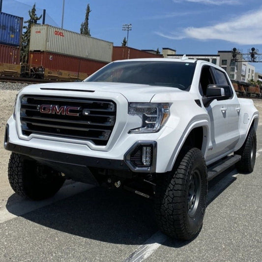 A large white GMC pickup truck with off-road modifications, including ADV Fiberglass 2019-2021 GMC Sierra fenders, is parked on a street. The vehicle boasts a rugged front bumper, lifted suspension, and large all-terrain tires. In the background, there are shipping containers and a section of a railroad track.