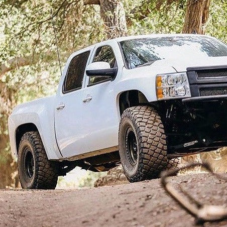 A white off-road truck with large, rugged tires is parked on a dirt trail in a forested area. Sunlight filters through the trees, highlighting the truck's elevated stance and sturdy body equipped with ADV Fiberglass 2007-2013 Chevrolet Silverado fenders. The surrounding foliage is dense, with green leaves and tall trees.