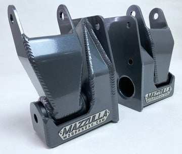 Displayed on a light surface are two gray metal bracket-like components, each with visible welds. These ruggedly designed components feature mounting holes, an angular structure, and a durable powder-coated finish. Engraved near the base is the brand name "MAZZULLA OFFROAD," ensuring lasting quality for your 1999-2018 CHEVY/GMC 1500 Shackle and Hanger kit.