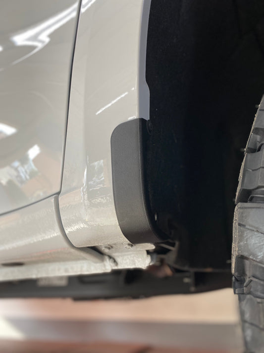 Close-up view of the rear fender area of a white Chevrolet Silverado 1500, focusing on the wheel well and part of the tire. The fender has a protective black trim near the edge. Noticeable is LTW Motorsports’ 2019-2024 Chevy Silverado 1500 Mud Flap Delete Kit, seamlessly integrated. The background is blurred, highlighting the vehicle detail.