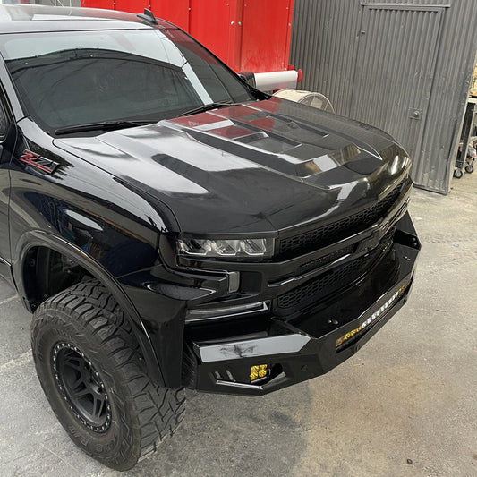 A black off-road truck with large, rugged tires and a stylish, aggressive front design is parked in a garage workshop. The ADV Fiberglass aftermarket hood for the 2019-2021 Chevrolet Silverado features sleek, angular lines, and there's a winch mounted on the front bumper. The garage surroundings have a mix of metal and red walls.