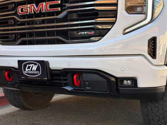 The front of a white GMC truck features red "GMC" grille lettering, a "LTW Motorsports" license plate, red tow hooks, and the newly installed 2022+ GMC Sierra 1500 Fog Light Kit with Baja Designs S2 Lights on the bumper.