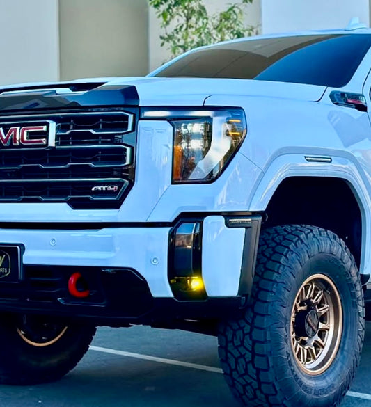 A front view of a white GMC AT4 truck with large off-road tires and black trim on concrete is enhanced by an LTW Motorsports 2022+ Chevy/GMC Truck Fog Light Kit with Baja Designs S1 Lights. A green leafy plant offers a serene backdrop.