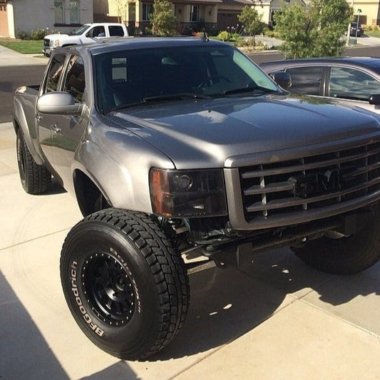 A silver-gray truck, likely one of the 2007-2013 GMC Sierras fitting ADV Fiberglass 2007-2013 GMC Sierra fenders, is parked in a residential driveway. The truck boasts large off-road tires and a lifted suspension. Part of another car is visible on the right, with houses and greenery in the background.
