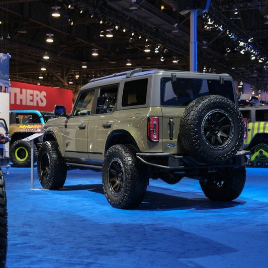 A tan-colored off-road vehicle with large, rugged tires, outfitted with the stylish ADV Fiberglass Rear Quarter Kit for the 2021-2023 Ford Bronco 4-Door, is showcased on a bright blue-carpeted floor in a dimly lit venue. The vehicle sports a spare tire mounted on the rear. Various other vehicles and large lights can be seen in the background.