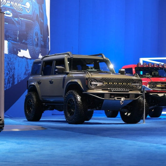 A gray Ford Bronco SUV equipped with ADV Fiberglass 2021-2023 Ford Bronco Fenders is displayed on a blue stage set against a bright blue backdrop. The stage also features a large screen presenting a close-up of the Bronco's front and another red Bronco parked in the background. The entire scene is well-lit, accentuating the vehicles.
