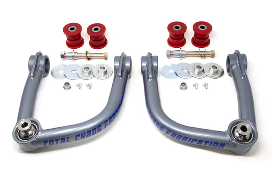 The 21-23 Ford Bronco Upper Control Arms by TOTAL CHAOS feature two metallic arms with red urethane bushings, uniballs, bolts, washers, and spacers displayed on a white surface.
